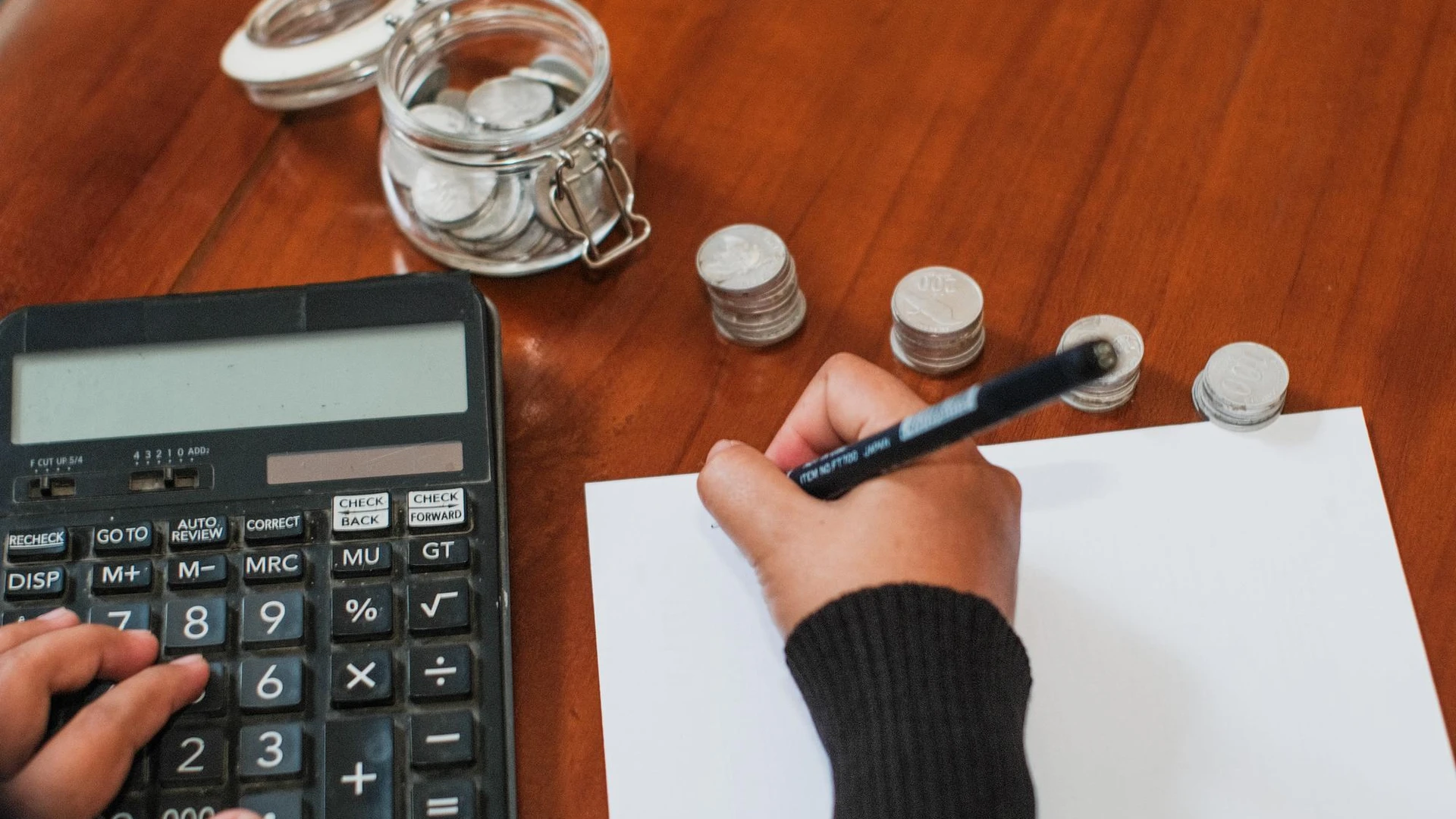 a person writing on a piece of paper with pen and calculator with stacks of coins on a wooden table